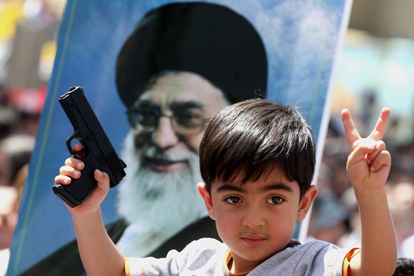 An Iraqi boy living in Iran holds a toy gun and flashes a victory sign in front of a poster of the Iranian Supreme leader Ayatollah Ali Khamenei in a demonstration against Sunni militants of the al-Qaida-inspired Islamic State of Iraq and the Levant, or ISIL, and to support the Grand Ayatollah Ali al-Sistani, Iraq's top Shiite cleric, in Tehran, Iran, Friday, June 20, 2014. (AP Photo/Ebrahim Noroozi)